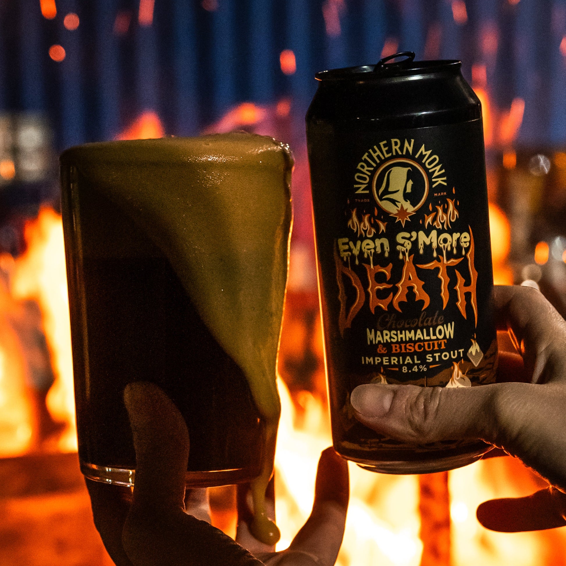 Two hands holding a glass of dark beer and a can of 'Even S'more Death' Imperial Stout in front of a fire.