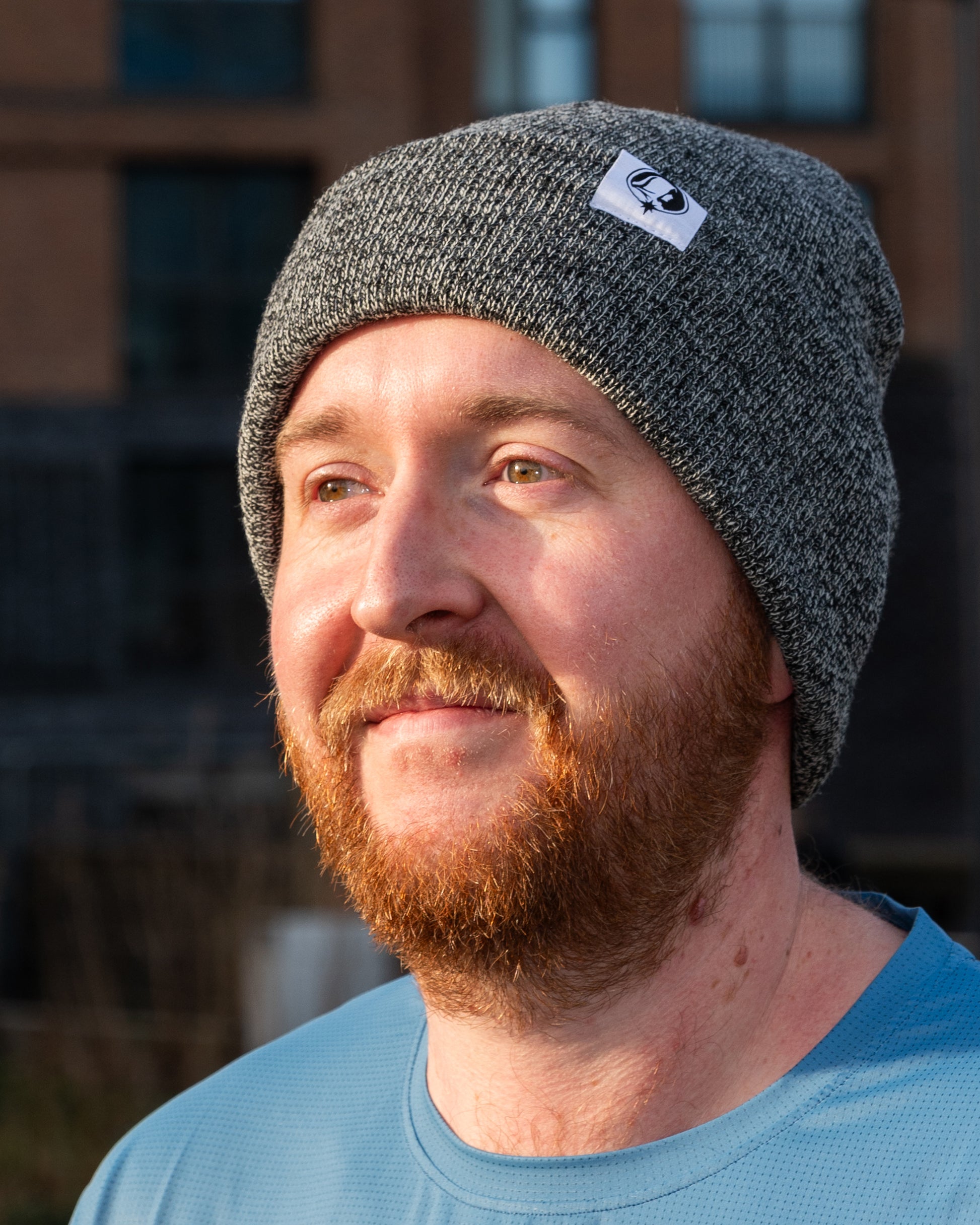 Man wearing a grey knit beanie with a Northern Monk logo tab, standing outdoors.