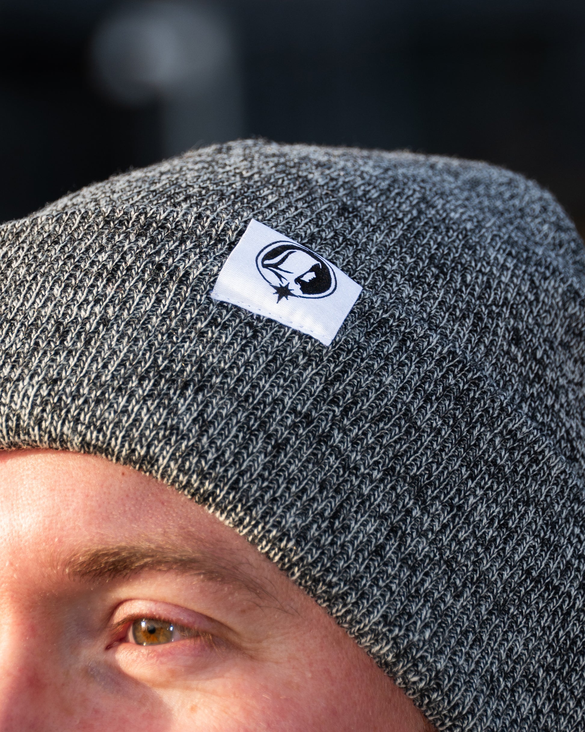 Person wearing a grey knit beanie with a Northern Monk logo tab on a blurred background